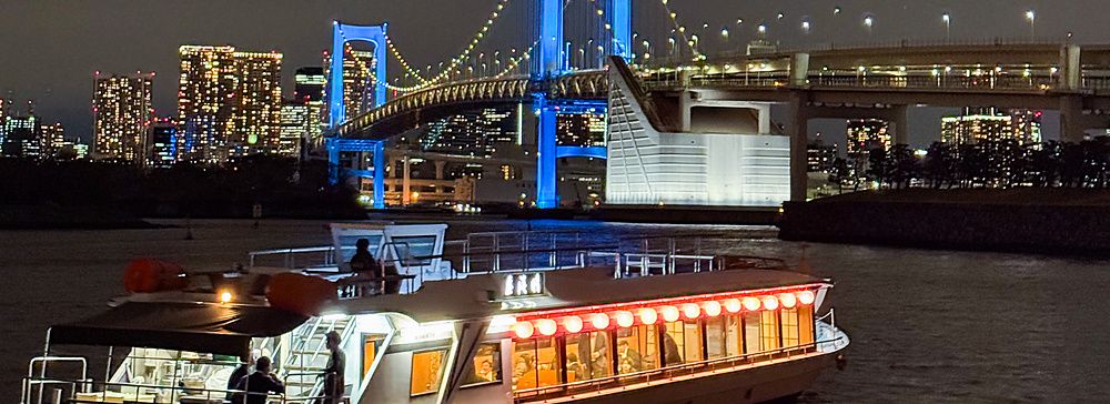 Veduta notturna di una Yakatabune che naviga vicino ad Odaiba, con il Rainbow Bridge illuminato sullo sfondo.