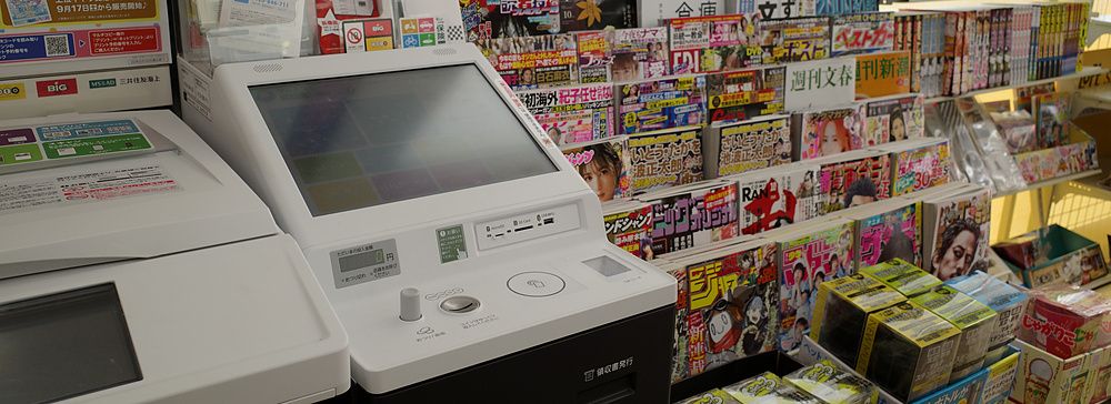 Postazione con stampante multifunzione e scaffali pieni di riviste in un minimarket a Tokyo.