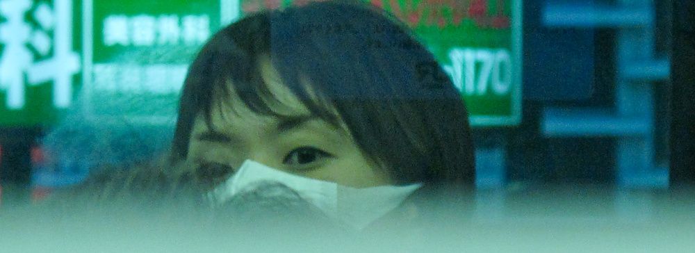 Persona con mascherina riflessa su vetro davanti a insegne luminose in Giappone.