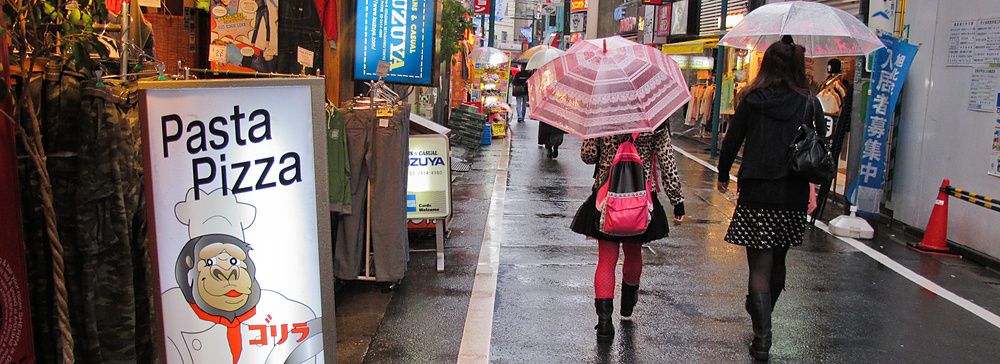 Strada piovosa a Shimokitazawa, Giappone, con insegna "Pasta Pizza" e persone con ombrelli.