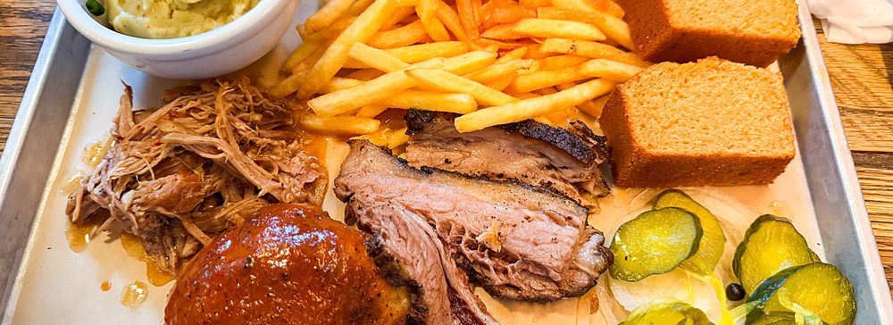 Un vassoio metallico di Smokehouse con barbecue, patatine fritte, pane di mais, purè e cetriolini.
