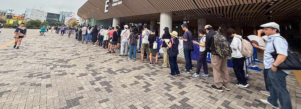 Una fila di persone in attesa all'ingresso dello Stadio Nazionale Giapponese a Tokyo su un piazzale pavimentato.