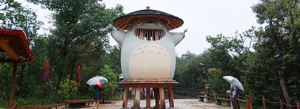 Grande statua in legno di Totoro in un parco boschivo sotto una pioggia leggera con visitatori.