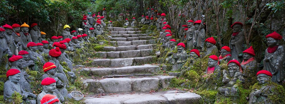 Molte statue di Buddha in pietra con cappelli rossi posizionate lungo una scalinata in un giardino a Miyajima.