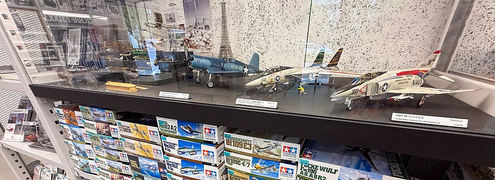 Scaffali pieni di scatole di modellini Tamiya sotto una vetrina con aerei assemblati al Tamiya PlaModel Factory a Tokyo.