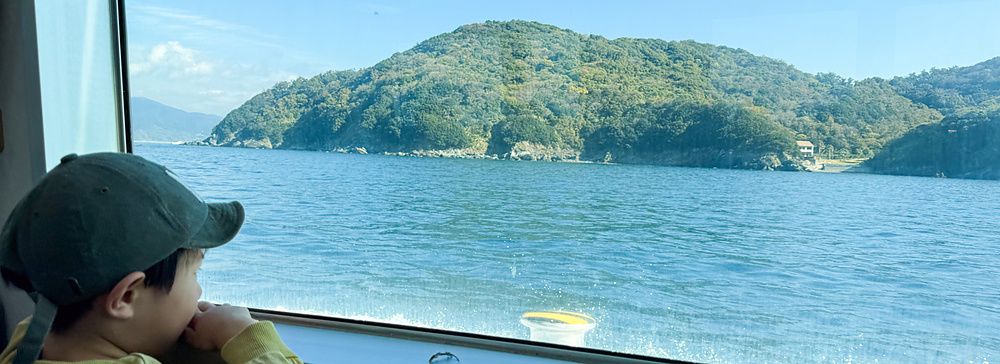 Un bambino guarda fuori dal finestrino di un traghetto per l'isola di Nushima verso la costa verdeggiante.