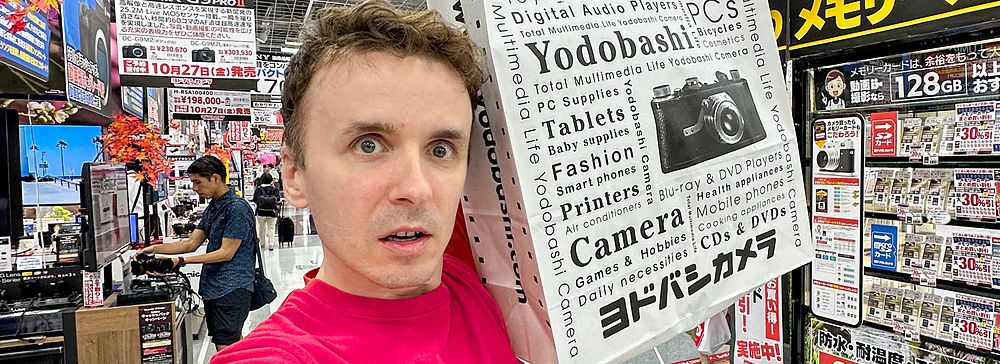 Marco Togni con busta di Yodobashi Camera in un negozio di elettronica.