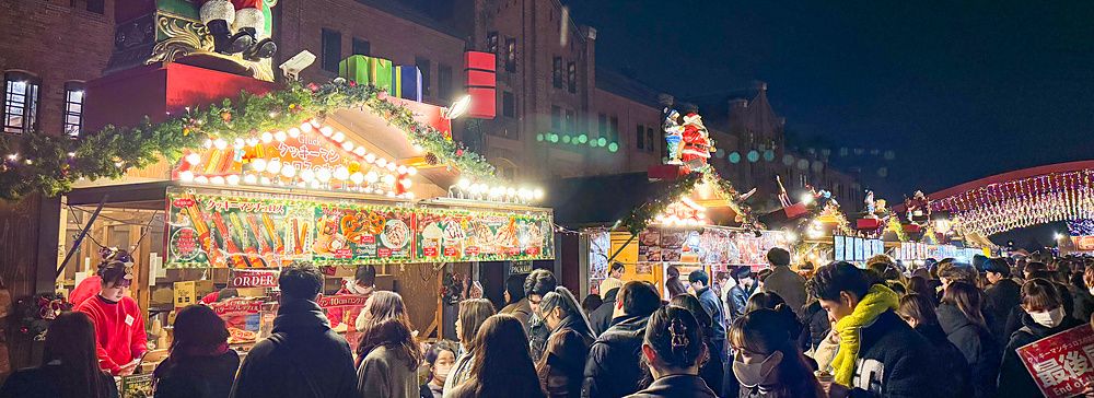 Veduta serale del Yokohama Red Brick Christmas Market con persone in coda davanti a chioschi decorati a tema natalizio.