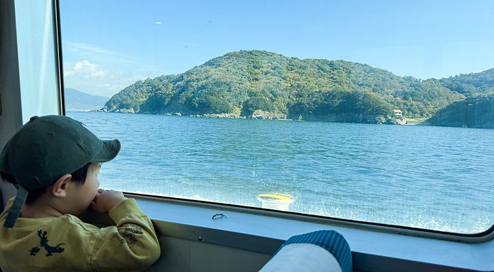 Un bambino guarda fuori dal finestrino di un traghetto per l'isola di Nushima verso la costa verdeggiante.