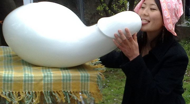 Una donna con fazzoletto rosa interagisce con una scultura bianca astratta su un tavolino all'aperto.