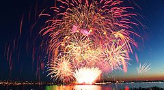 Spettacolo di fuochi d'artificio colorati riflessi sull'acqua al tramonto.