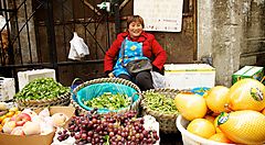 Venditrice di frutta e verdura al mercato seduta tra ceste di meloni, uva e verdure.