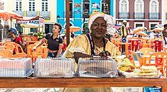 Donna in abiti tradizionali brasiliani con cibo su un banco in una piazza colorata.