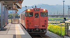 Treno rosso in una stazione di Takaoka, Giappone, con monti sullo sfondo.
