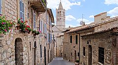 Strada acciottolata di Assisi con edifici in pietra e campanile sullo sfondo.