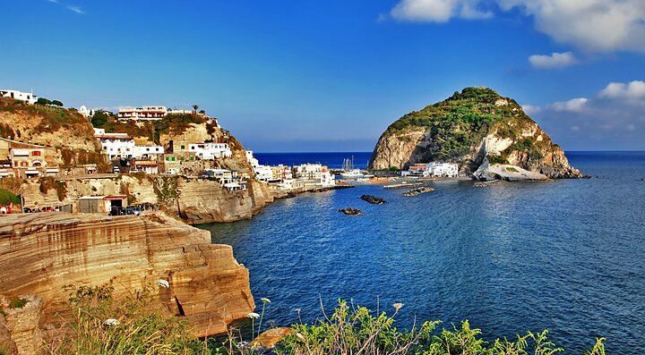 Vista panoramica del porto di Ischia con mare cristallino e colline verdeggianti.