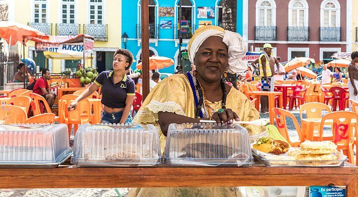 Donna in abiti tradizionali brasiliani con cibo su un banco in una piazza colorata.
