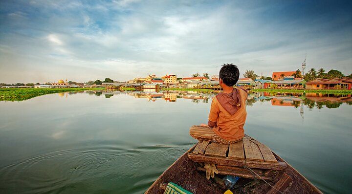 Bambino su una barca in un fiume in Cambogia, villaggio sullo sfondo.