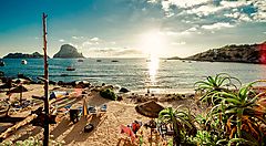 Tramonto su una spiaggia di Ibiza con mare, palme e barche all'orizzonte.