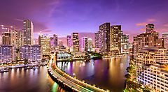 Skyline di Miami al tramonto con grattacieli illuminati e fiume in primo piano.