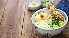 Una ciotola di Sanuki Udon con tempura e cipollotto su tavolo di legno.