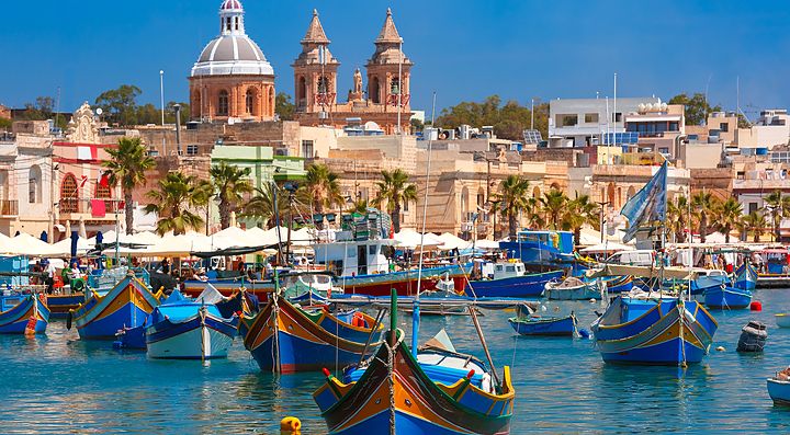 Porto colorato a Malta con barche tradizionali e edifici storici sullo sfondo.