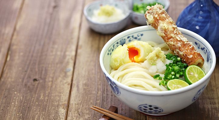 Una ciotola di Sanuki Udon con tempura e cipollotto su tavolo di legno.