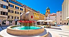 Fontana in una piazza di Fiume con edifici storici sullo sfondo.