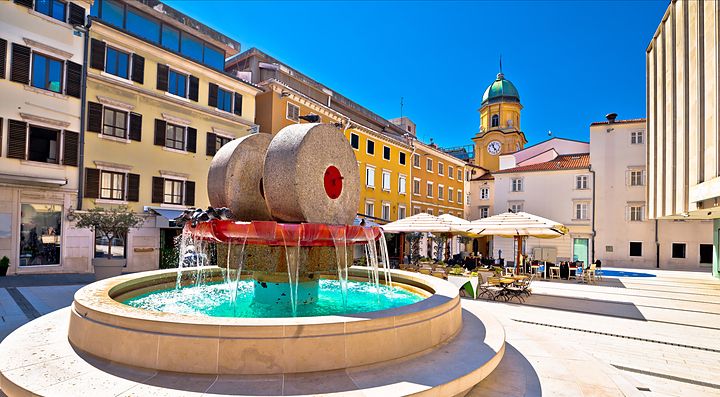 Fontana in una piazza di Fiume con edifici storici sullo sfondo.