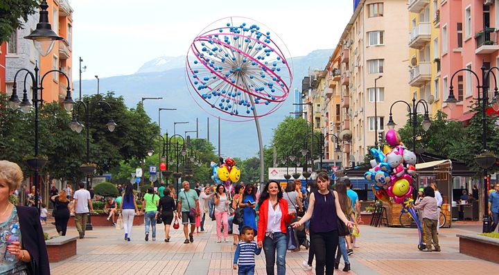 Strada pedonale affollata a Sofia con monumento colorato al centro.