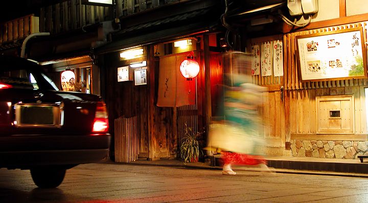 Strada notturna nel quartiere di Gion, Kyoto, con lanterne e figure in movimento.