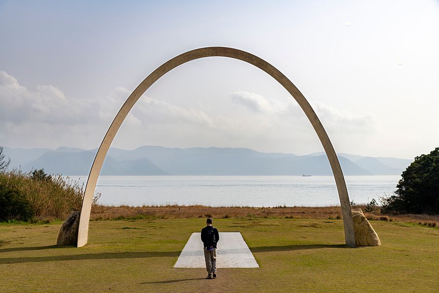 Una persona cammina verso un grande arco architettonico in cemento posizionato su un prato affacciato sul mare.