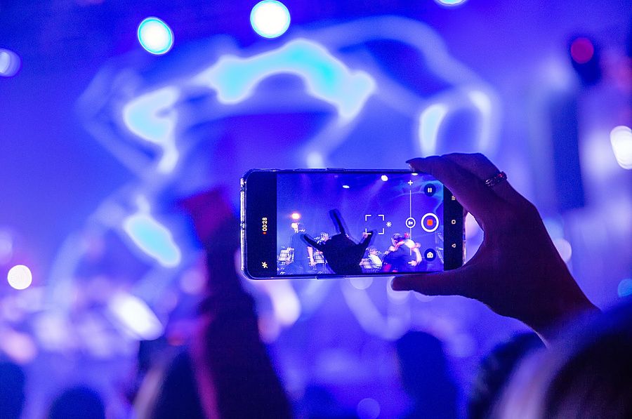Primo piano di una mano che usa uno smartphone per filmare un concerto con luci blu intense sullo sfondo.