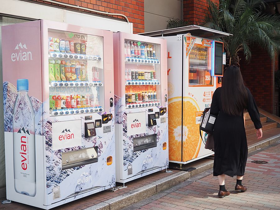 Una persona cammina accanto a tre distributori automatici di bevande decorati con grafiche Evian e agrumi a Tokyo.