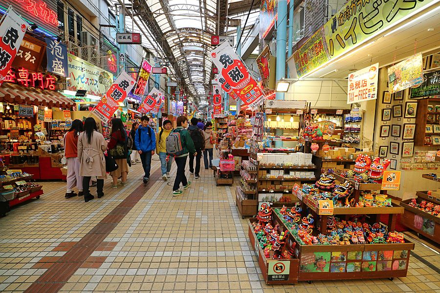 Galleria coperta Heiwa Dori a Naha, Okinawa, con negozi di souvenir, insegne colorate e passanti che camminano.