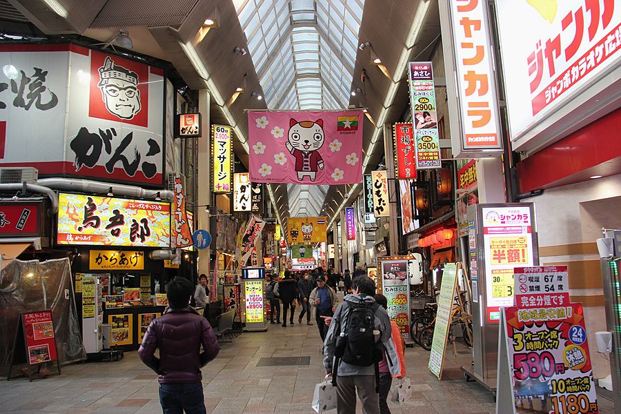 Persone che camminano lungo la via commerciale coperta di Shinsaibashi a Osaka con insegne luminose e stendardi colorati.