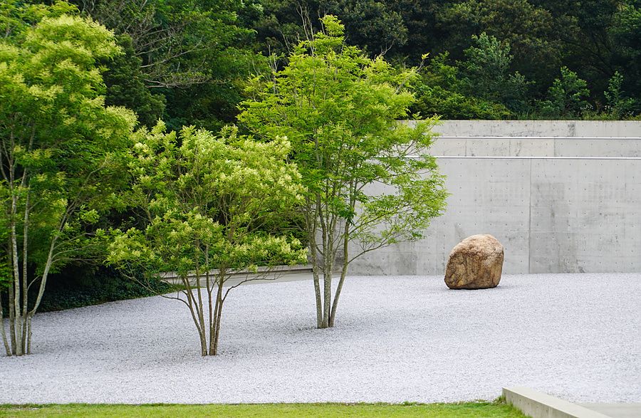 Giardino zen con alberi e una roccia solitaria su ghiaia chiara davanti a una parete in cemento al Lee Ufan Museum.