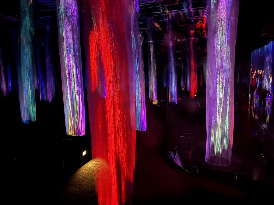 Installazione artistica con cilindri luminosi sospesi in uno spazio buio e riflettente.