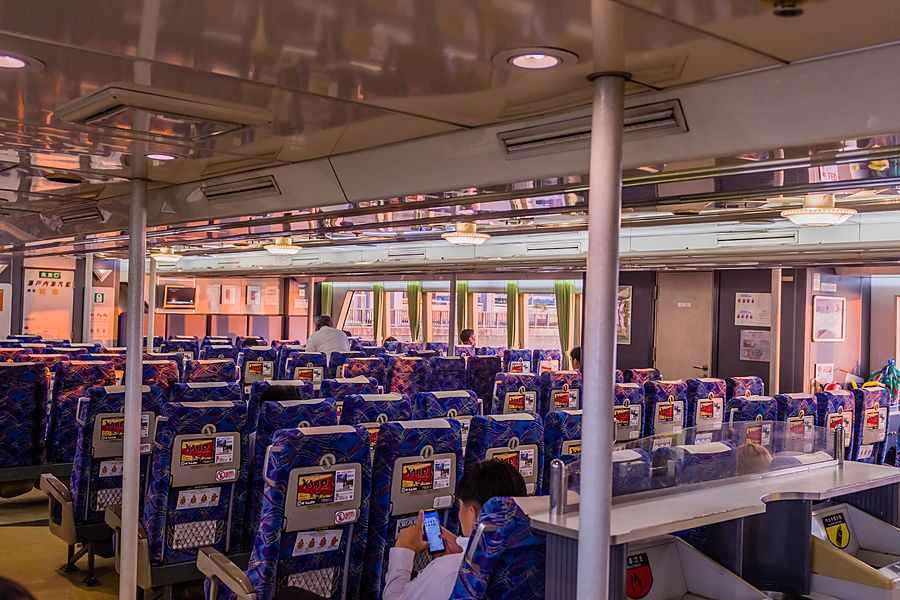 Interno del ponte inferiore di un traghetto con sedili blu e persone sedute a Matsuyama, Giappone.