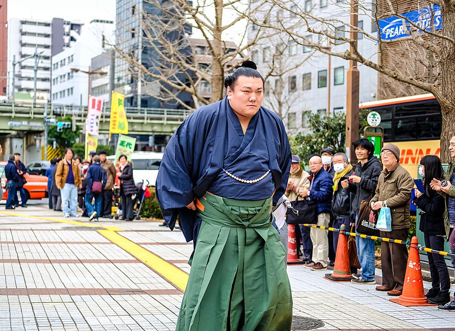 Lottatore di sumo in kimono blu e hakama verde cammina per strada a Tokyo tra le persone.
