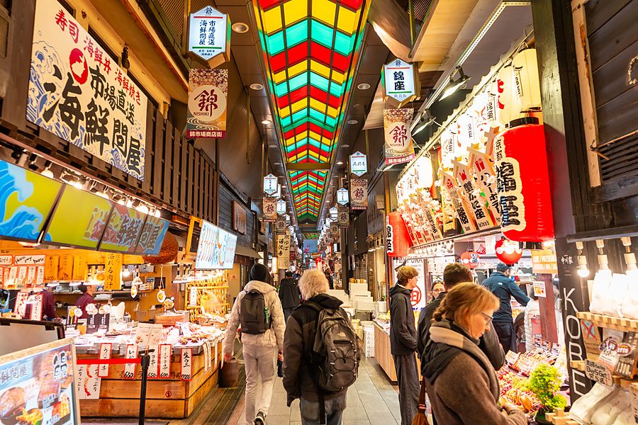 Persone camminano sotto il tetto colorato del mercato coperto Nishiki a Kyoto tra bancarelle di cibo tipico.