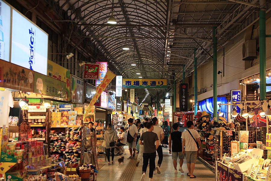 Corridoio coperto di un mercato pubblico a Naha con negozi, turisti e insegne luminose in giapponese.