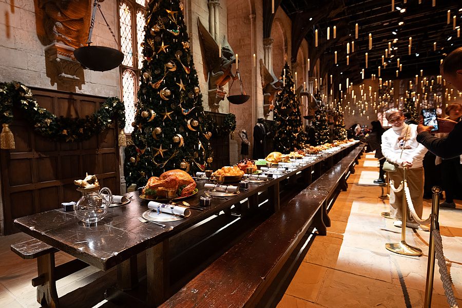 Lunga tavola di legno imbandita per un banchetto natalizio con alberi decorati e candele sospese in una grande sala.