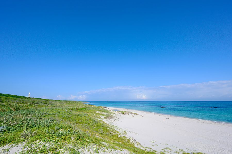 Veduta panoramica di una spiaggia bianca con mare turchese e un piccolo faro in lontananza.
