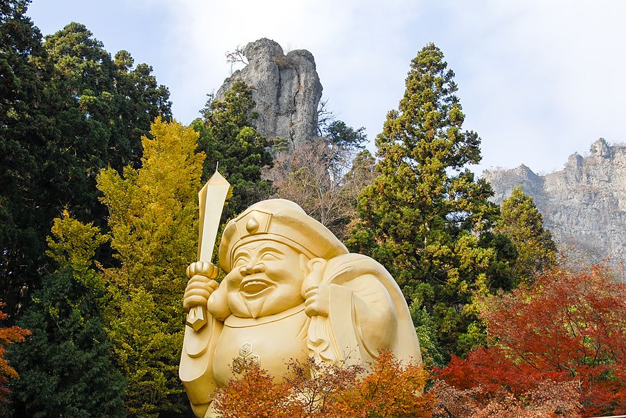 Grande statua dorata di Daikokuten circondata da alberi autunnali e rocce scoscese al santuario Nakanotake in Giappone.