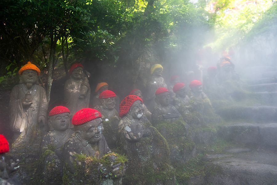 Piccole statue di Jizo in pietra con cappellini rossi allineate su un sentiero boscoso nebbioso al tempio Daisho-in.