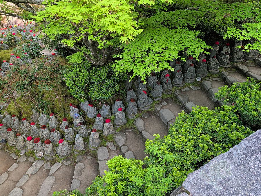 Piccole statue Jizo con cappelli rossi lungo un sentiero di pietra in un giardino giapponese sotto alberi di acero.
