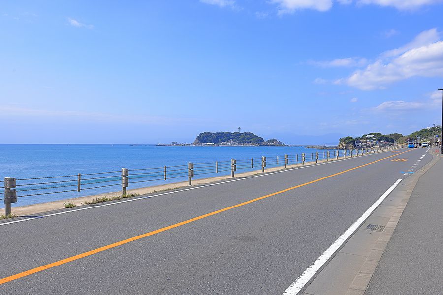 Strada costiera asfaltata che costeggia il mare con vista sull'isola di Enoshima in una giornata serena.