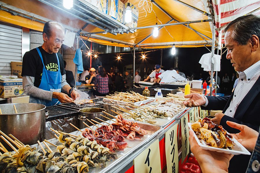 Un uomo griglia spiedini di carne in una bancarella di un mercato notturno a Tokyo mentre un cliente attende il cibo.