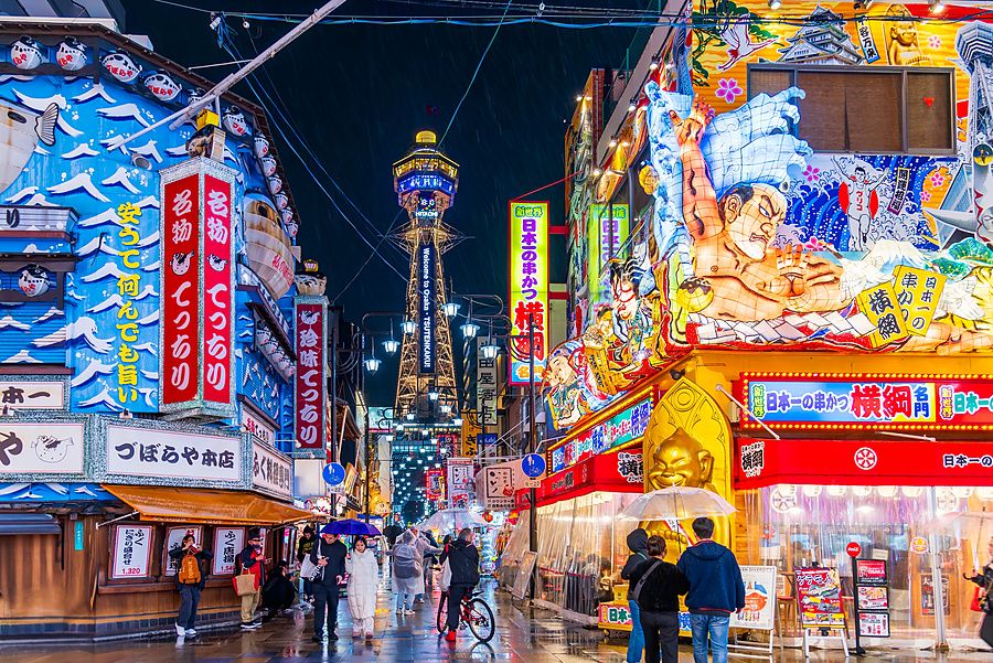 Veduta notturna e piovosa di una strada nel quartiere Shinsekai a Osaka con la torre Tsutenkaku illuminata sullo sfondo.